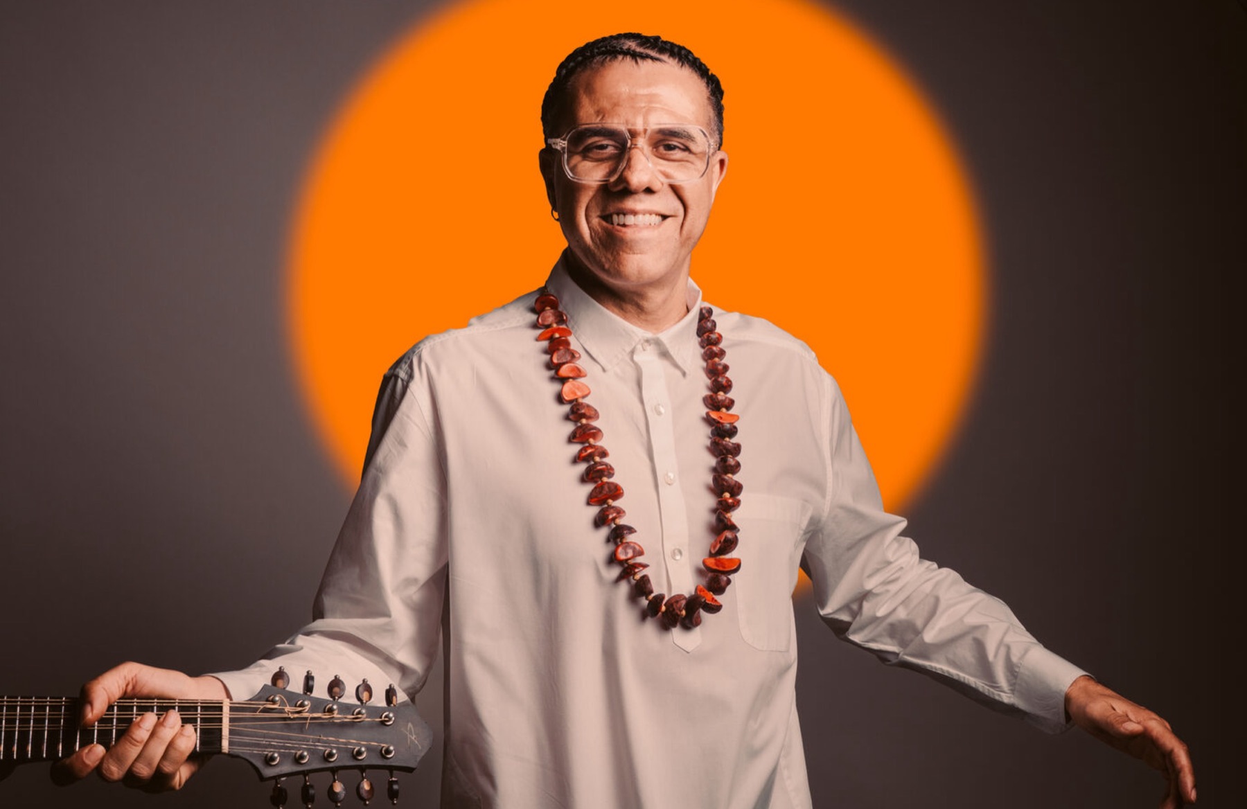 Hamilton de Holanda sorrindo, de camisa branca e colar de sementes, diante de um círculo laranja