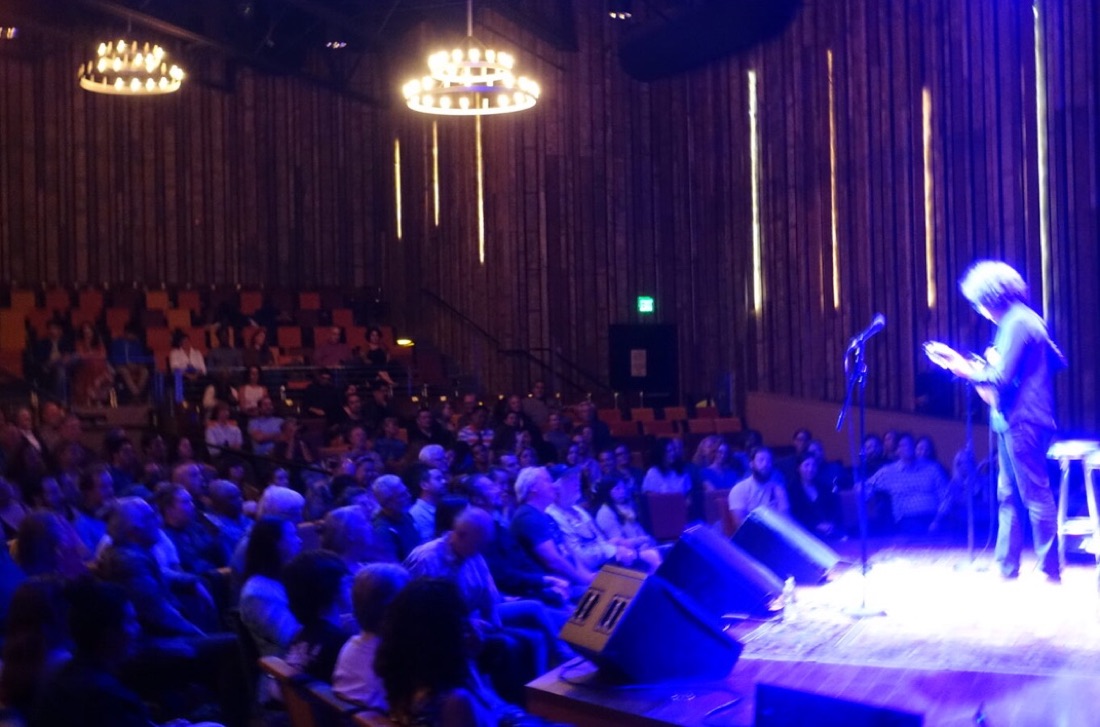 Hamilton de Holanda no palco de um auditório, sob luz azul, diante do público